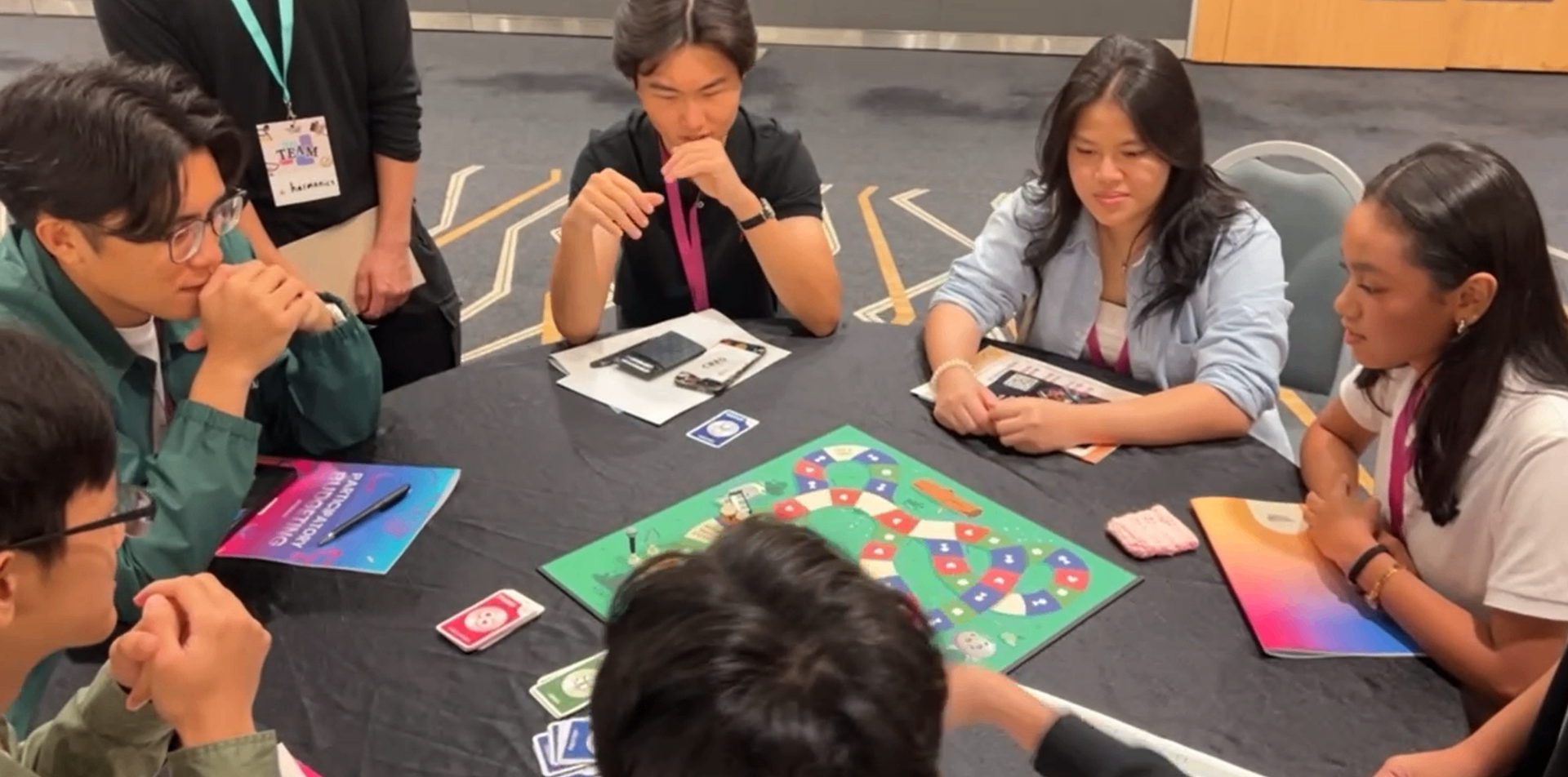 A group of players gathered around the Harmony Quest board during a playtesting session.